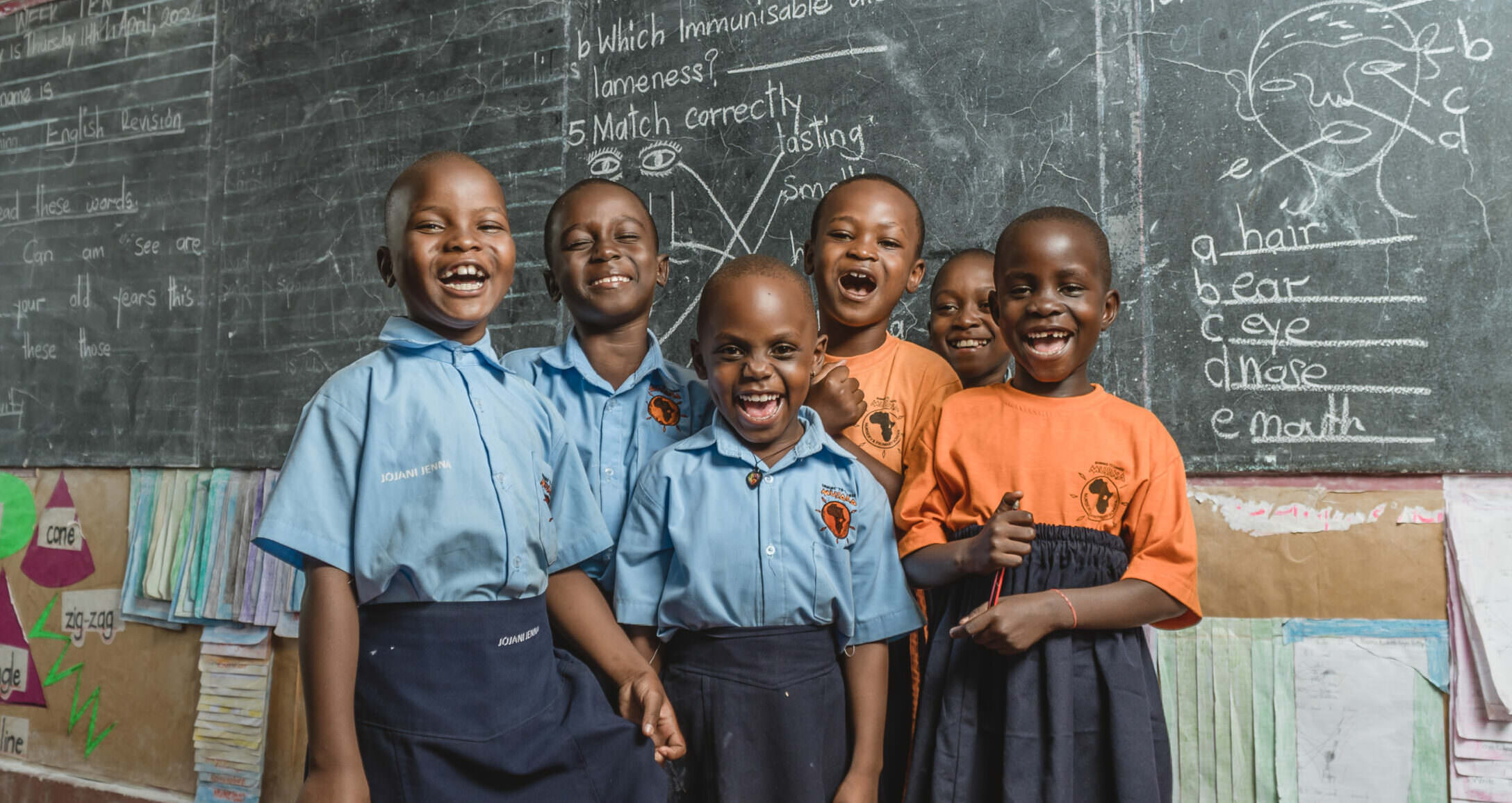Group of smiling Musana Primary School students standing in front of a chalkboard filled with lesson content and educational diagrams.