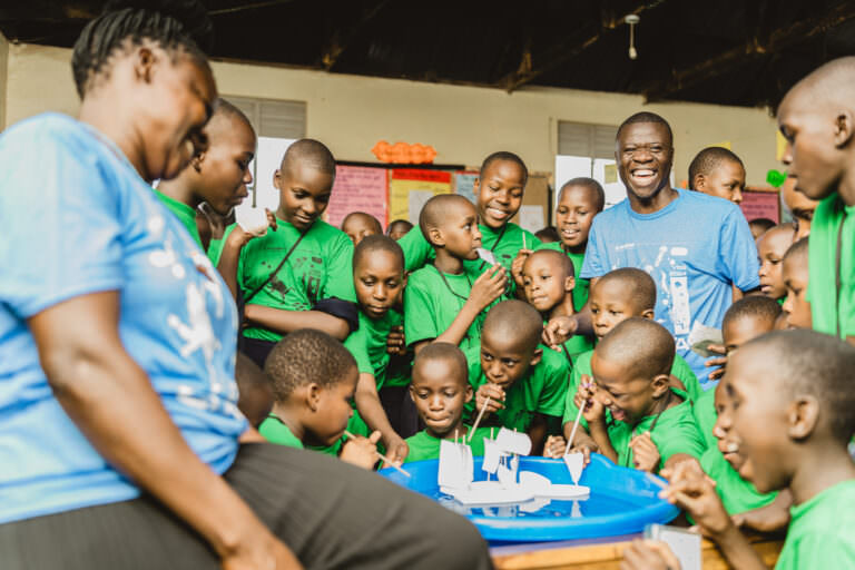 A group of smiling schoolchildren in green uniforms gathered around a blue basin with floating paper boats, participating in a hands-on activity. Two adult facilitators, one in a blue shirt, engage with the children in a brightly decorated classroom