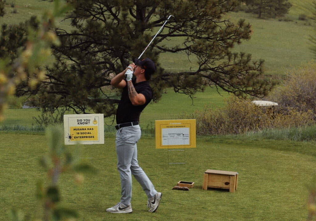 A golfer tees off at the Musana Annual Golf Tournament at Colorado Golf Club, with signs in the background highlighting Musana’s social enterprises.