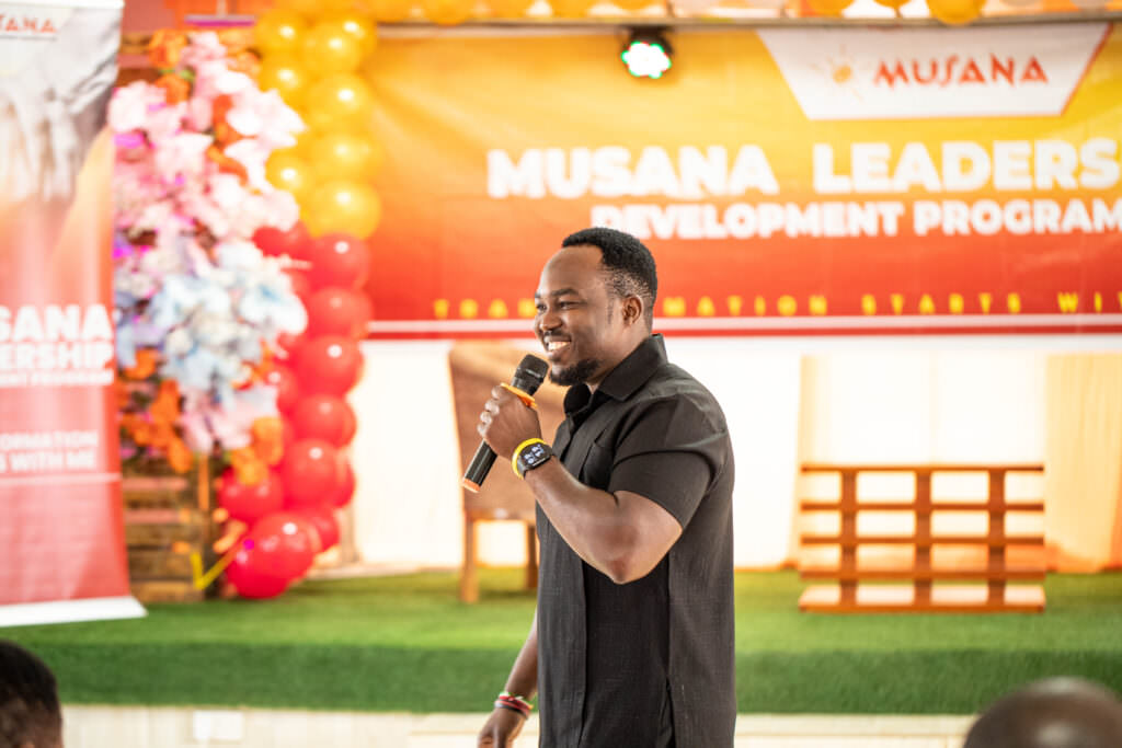 Haril, a Musana leader, smiles while speaking into a microphone on stage during the Musana Leadership Development Program. The background features colorful balloons and a vibrant banner with the Musana logo.